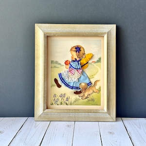 May include: Framed vintage print featuring a young girl in a blue dress, holding an apple and a yellow hat, running with a small dog. The artwork has a light-colored wooden frame and depicts a pastoral scene with flowers and a path.
