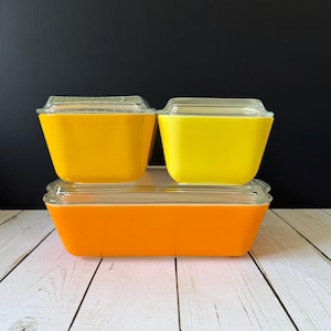 May include: Three vintage Pyrex food storage containers. Two square containers in yellow and one rectangular container in orange, all with clear glass lids. The containers are stacked on a white wooden surface against a black background.