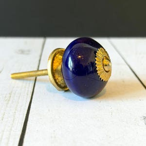 May include: A round, deep blue ceramic cabinet knob with a glossy finish. The knob features a gold-toned metal base and a decorative, scalloped gold-toned metal plate at the front center, attached to a threaded metal bolt for installation.