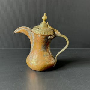 May include: A vintage copper and brass coffee pot with a curved spout and a handle. The pot has a decorative lid and a bulbous body. The metal has a weathered appearance with areas of oxidation, giving it an antique look. The pot is likely used for serving coffee or tea.