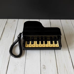 Puede incluir: Un teléfono negro con forma de piano de cola. El teléfono tiene un cuerpo negro con teclas amarillas y negras, y un cable de auricular en espiral. Los números 1-0 están impresos en las teclas.