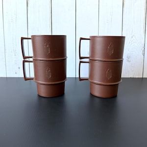 May include: Four brown stackable mugs with angular handles. Each mug features a small, embossed leaf-shaped design on the front. The mugs are arranged in two vertical stacks of two, showcasing a uniform matte finish and a simple, functional design.
