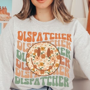 May include: A gray sweatshirt with a retro-style smiley face graphic and the word "DISPATCHER" repeated in a colorful, layered design.