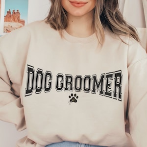 May include: A beige sweatshirt with the text "DOG GROOMER" in black block letters with a paw print below.