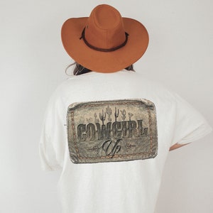 May include: White t-shirt with a brown leather patch on the back that says "Cowgirl Up" with a cactus design. The shirt is being worn by a person wearing a brown cowboy hat.