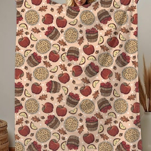 May include: A beige blanket with a repeating pattern of red apples, apple slices, pie, and barrels. The blanket is held up, showcasing the fall-themed design. The blanket is a cozy home decor item.