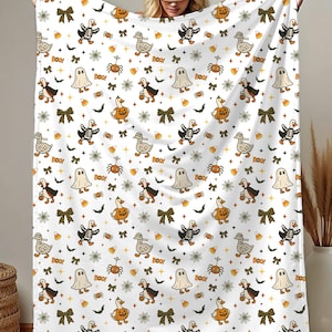 May include: White Halloween-themed blanket featuring a repeating pattern of cartoon geese, skeletons, ghosts, pumpkins, spiders, bows, and the word "BOO!" in orange and black. The blanket is held up by a person.