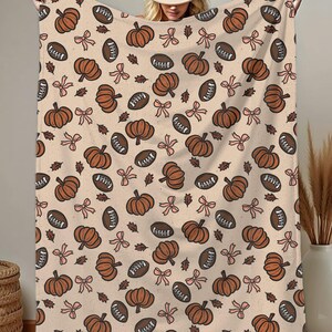 May include: A beige blanket with a repeating pattern of brown pumpkins, footballs, bows, and leaves. The blanket is held up, showcasing the fall-themed design. The blanket is made of a soft material.