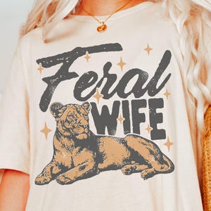 May include: A white t-shirt with the text "Feral Wife" in black lettering and a brown and black lion illustration. The shirt has a distressed vintage look.