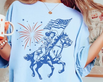 May include: A light blue t-shirt with a vintage graphic of a woman riding a horse, holding an American flag. Fireworks and stars are in the background. The text "Chambray" is in the top left corner.