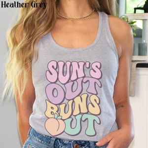 May include: A heather grey tank top with the text "SUN'S OUT BUNS OUT" in a retro font. The text is in pink, purple, yellow, and green. A peach graphic is below the text.