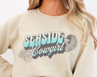May include: A light beige sweatshirt with the text "Seashore Cowgirl" in blue and gray lettering with a seashell design.