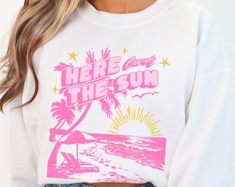 May include: White sweatshirt with a pink graphic design of palm trees, a beach scene, and the text "Here Comes The Sun".
