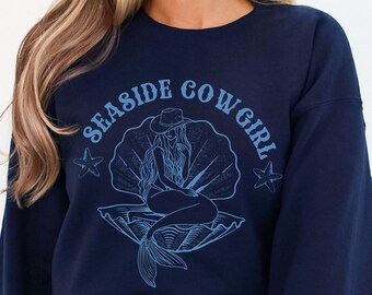 May include: Navy blue sweatshirt with a graphic of a mermaid wearing a cowboy hat and sitting in a seashell. The text "SEASIDE COWGIRL" is printed in a retro font around the graphic.