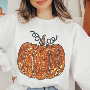 Puede incluir: Sudadera blanca con un diseño de calabaza naranja brillante. La calabaza tiene un tallo y enredaderas verdes.