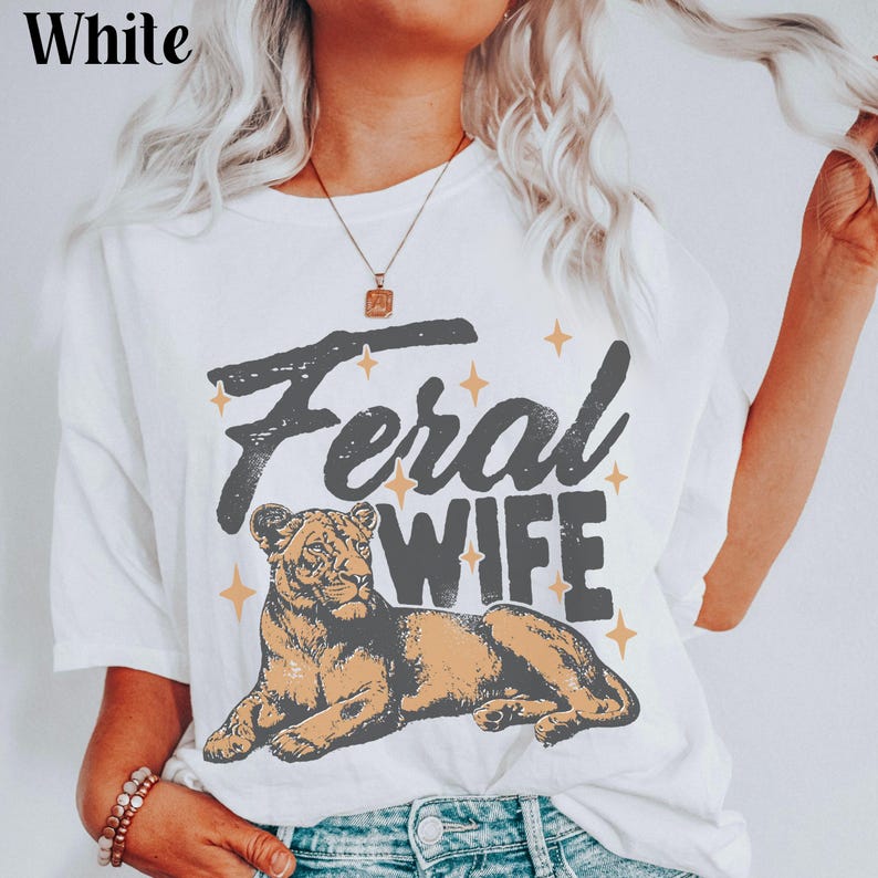 May include: White t-shirt with a graphic of a lioness and the text "Feral Wife" in black lettering with gold stars.