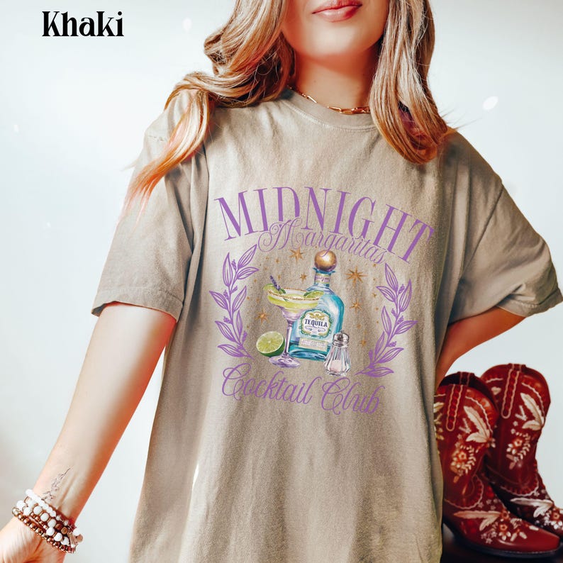 Pu&ograve; includere: Una t-shirt color kaki con una stampa grafica di un cocktail margarita con il testo "Midnight Margarita Cocktail Club" in lettere viola.