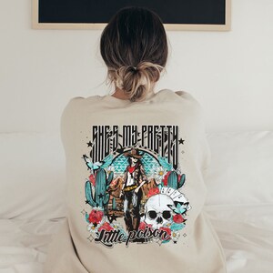 May include: A beige sweatshirt with a graphic print of a woman in a cowboy hat and a skull. The text "She's my pretty little poison" is printed on the sweatshirt.