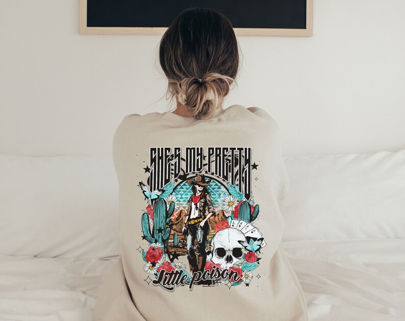 May include: A beige sweatshirt with a graphic print of a woman in a cowboy hat and a skull. The text "She's my pretty little poison" is printed on the sweatshirt.