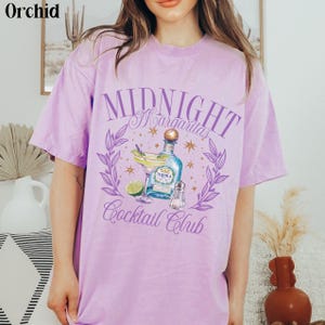 Pu&ograve; includere: Maglietta viola con un design grafico di un cocktail margarita, una bottiglia di tequila e il testo "Midnight Margaritas Cocktail Club".