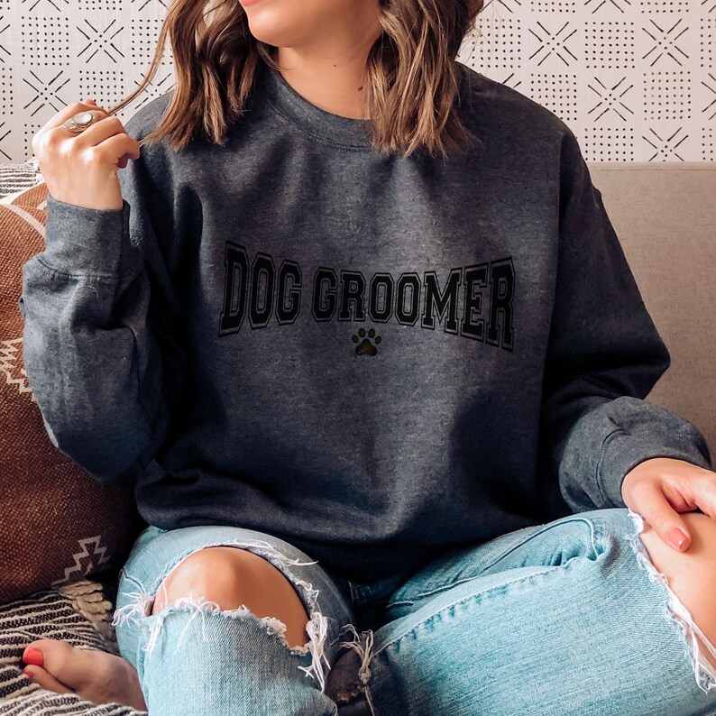 May include: A dark gray sweatshirt with the text "DOG GROOMER" in black letters with a paw print in the center. The model is wearing blue jeans.