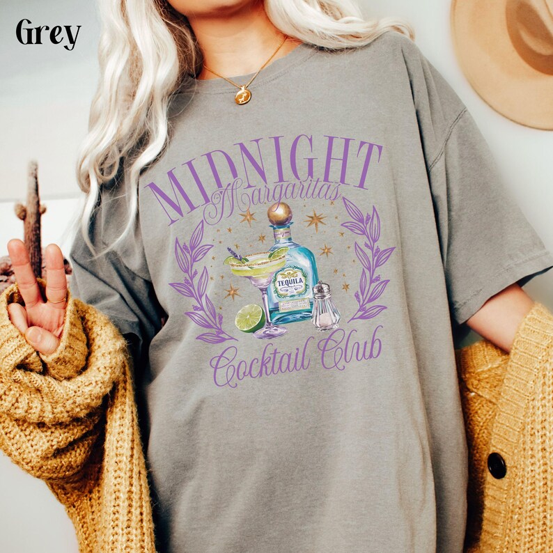 Pu&ograve; includere: Una maglietta grigia con un design grafico viola che dice "Midnight Margaritas Cocktail Club". Il grafico presenta una bottiglia di tequila, un bicchiere di margarita, un lime e un saliera.