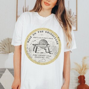 May include: White t-shirt with a yellow circle graphic featuring a vintage illustration of a grindstone with the text "Nose on the Grindstone Don't Lie & Don't Steal Pike County Coal Kentucky, USA"