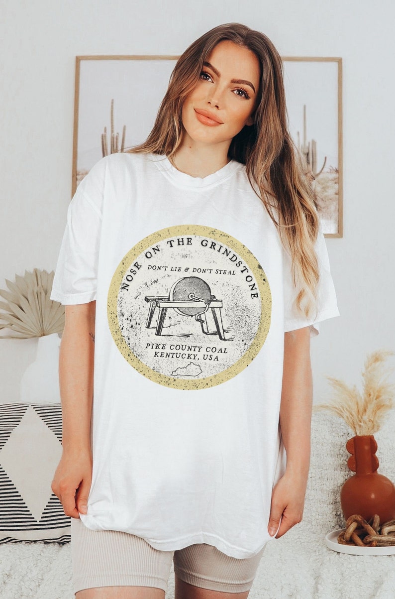 May include: White t-shirt with a yellow circle graphic featuring a vintage illustration of a grindstone with the text "Nose on the Grindstone Don't Lie & Don't Steal Pike County Coal Kentucky, USA"