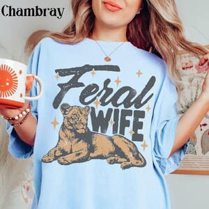 May include: A light blue t-shirt with a graphic of a lioness and the text "Feral Wife" in a vintage style font.