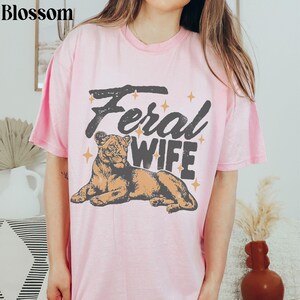 May include: Pink t-shirt with the text "Feral Wife" in a vintage style font. A lioness illustration is below the text.