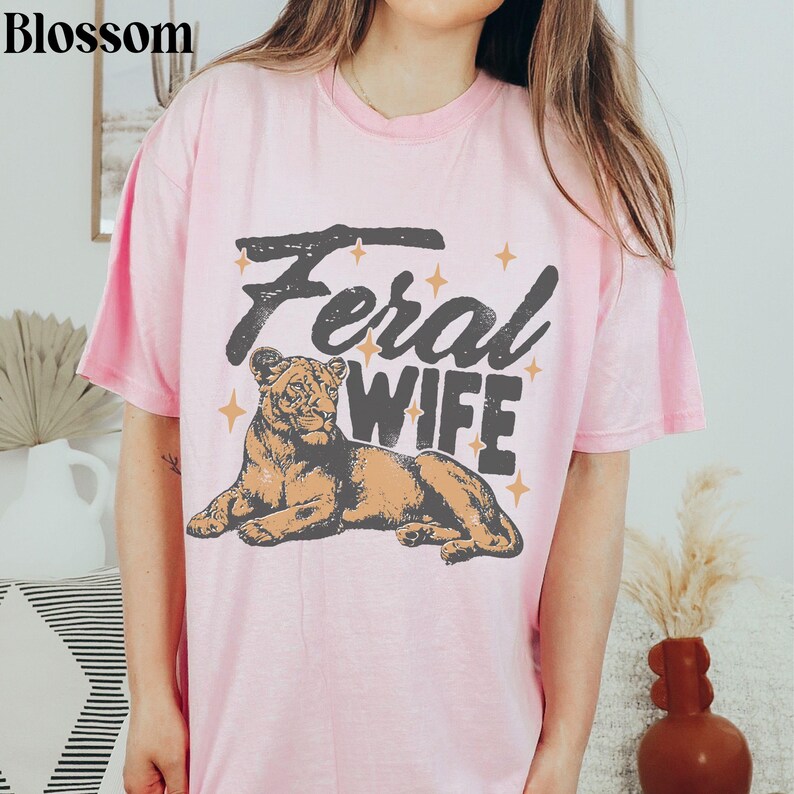 May include: Pink t-shirt with the text "Feral Wife" in a vintage style font. A lioness illustration is below the text.