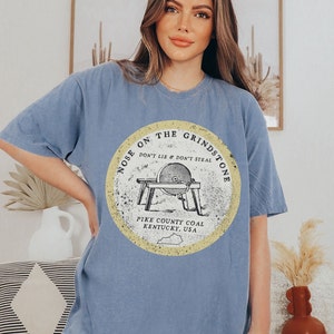 May include: A light blue t-shirt with a vintage-style graphic of a grindstone with the text "Nose on the Grindstone Don't Lie & Don't Steal Pike County Coal Kentucky, USA".