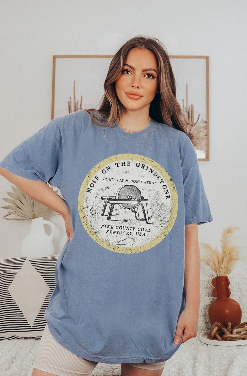 May include: A light blue t-shirt with a vintage-style graphic of a grindstone with the text "Nose on the Grindstone Don't Lie & Don't Steal Pike County Coal Kentucky, USA".