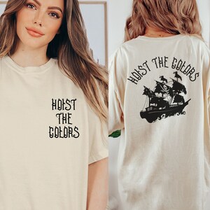 May include: A cream colored t-shirt with the text "Hoist the Colors" on the front and a black silhouette of a pirate ship with the text "Hoist the Colors" on the back.