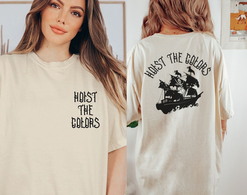 May include: A cream colored t-shirt with the text "Hoist the Colors" on the front and a black silhouette of a pirate ship with the text "Hoist the Colors" on the back.