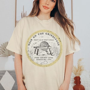 May include: A beige t-shirt with a vintage-style graphic of a grindstone with the text "Nose on the Grindstone Don't Lie & Don't Steal Pike County Coal Kentucky, USA".