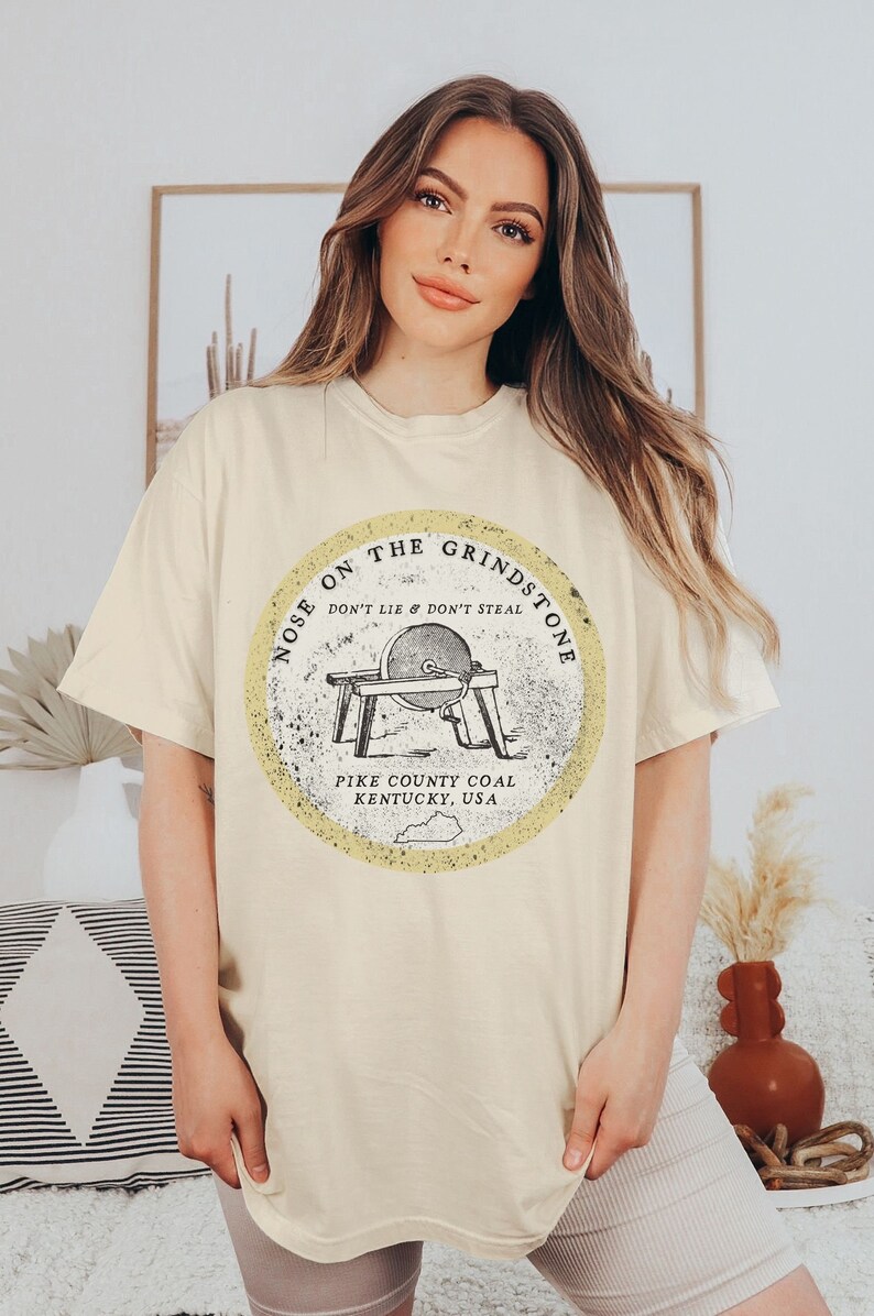 May include: A beige t-shirt with a vintage-style graphic of a grindstone with the text "Nose on the Grindstone Don't Lie & Don't Steal Pike County Coal Kentucky, USA".