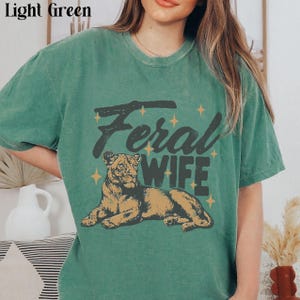 May include: A light green t-shirt with a graphic print of a lioness and the text "Feral Wife".