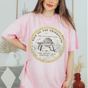 May include: A pink t-shirt with a graphic of a vintage grindstone with the text "Nose on the Grindstone Don't Lie & Don't Steal Pike County Coal Kentucky, USA".