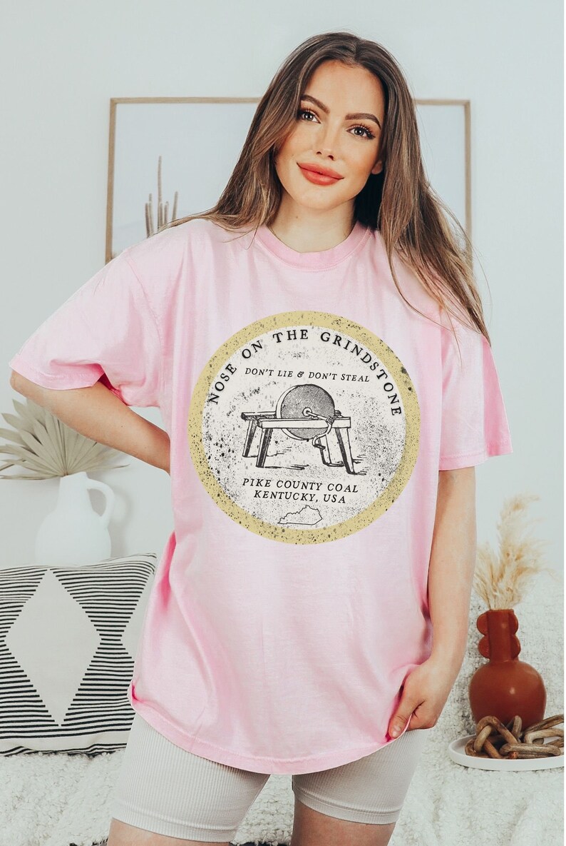 May include: A pink t-shirt with a graphic of a vintage grindstone with the text "Nose on the Grindstone Don't Lie & Don't Steal Pike County Coal Kentucky, USA".