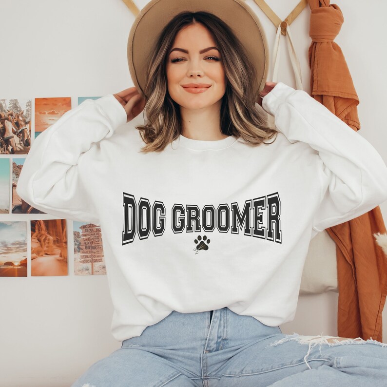 May include: A white sweatshirt with the text "DOG GROOMER" in black block letters with a paw print below. The model is wearing the sweatshirt and has brown hair and is wearing a brown hat.