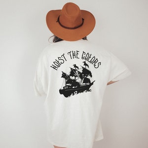 May include: White t-shirt with a black silhouette of a pirate ship and the text "Hoist the Colors" on the back.
