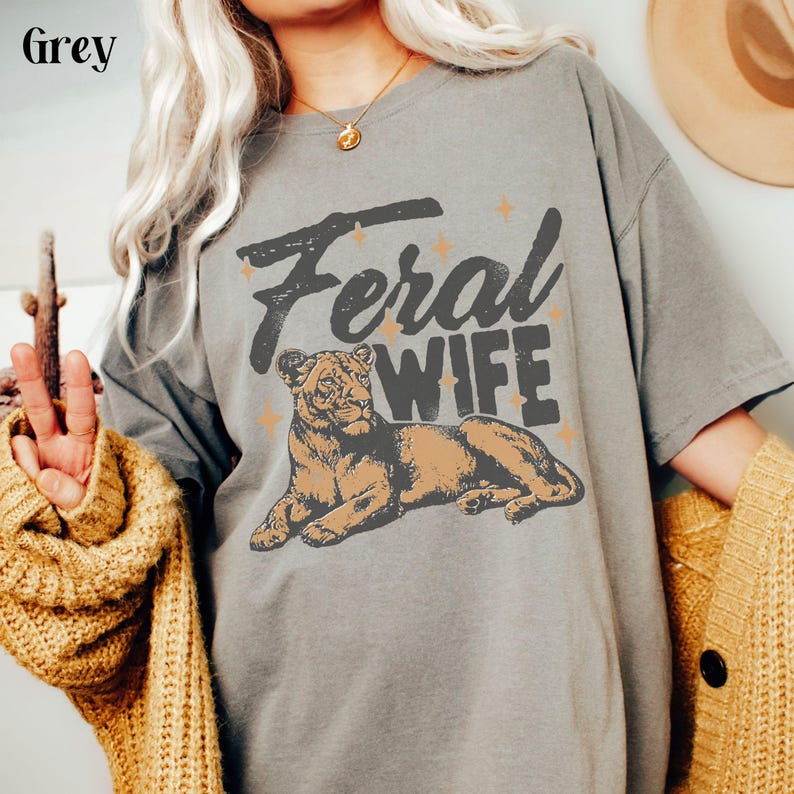 May include: A grey t-shirt with a graphic of a lioness and the text "Feral Wife" in black lettering.