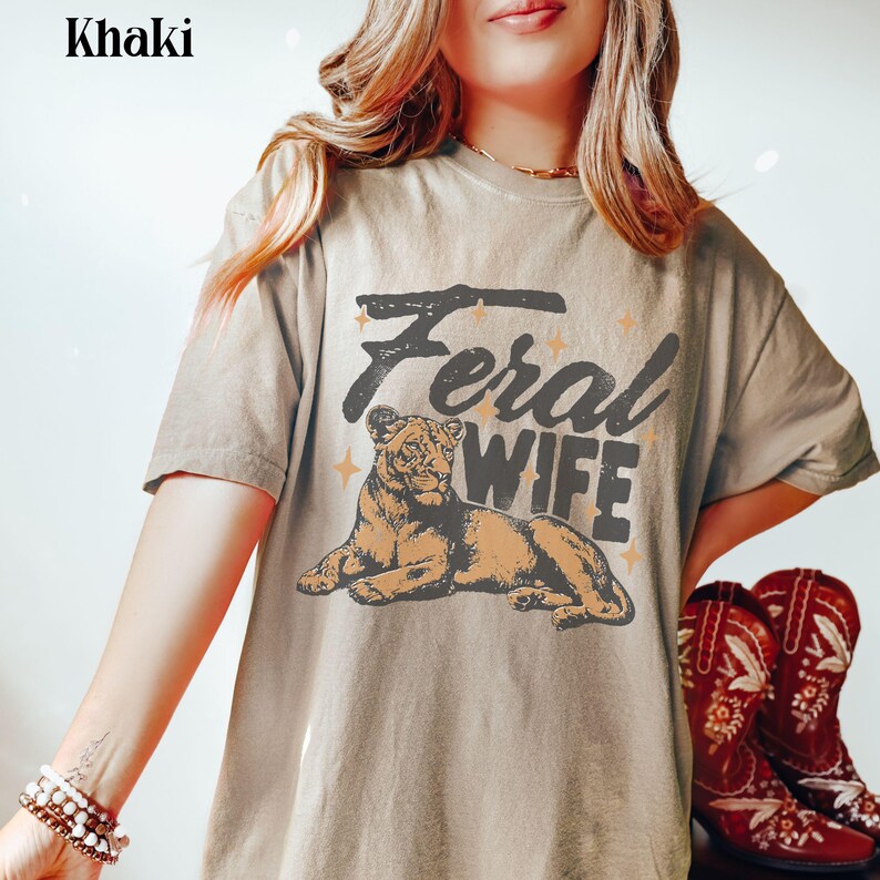 May include: A khaki t-shirt with a graphic of a lioness and the text "Feral Wife".