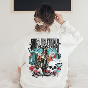 May include: White sweatshirt with a graphic print of a woman in a cowboy hat and a skull with flowers and cacti. The text "She's my pretty little poison" is printed below the graphic.