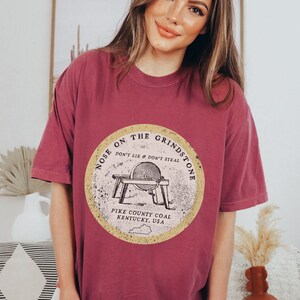 May include: A maroon t-shirt with a vintage-style graphic of a grindstone with the text "Nose on the Grindstone Don't Lie & Don't Steal Pike County Coal Kentucky, USA".