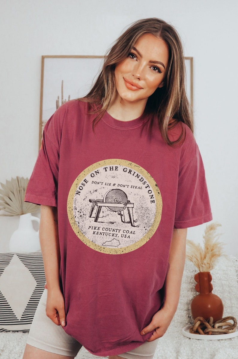 May include: A maroon t-shirt with a vintage-style graphic of a grindstone with the text "Nose on the Grindstone Don't Lie & Don't Steal Pike County Coal Kentucky, USA".