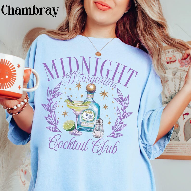 Pu&ograve; includere: Una maglietta blu chiaro con un design grafico viola che dice "Midnight Margaritas Cocktail Club". La grafica presenta una bottiglia di tequila, un bicchiere di margarita, un lime e un saliera. Il design &egrave; circondato da foglie viola e stelle dorate.