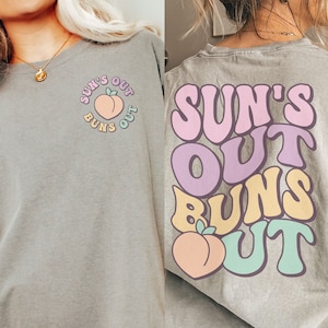 May include: A gray t-shirt with the text "Sun's Out Buns Out" in a retro font. The text is in pink, purple, yellow, and green. A peach graphic is below the text.