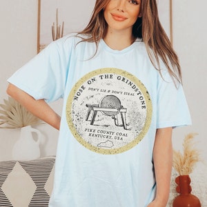 May include: Light blue t-shirt with a vintage-style graphic of a grindstone with the text "Nose on the Grindstone Don't Lie & Don't Steal Pike County Coal Kentucky, USA."